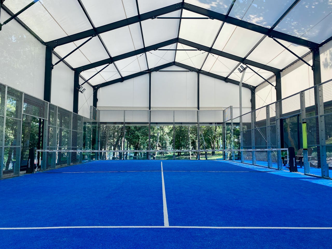about-us-02 Spacious indoor padel court with vibrant blue flooring under a wide roof.